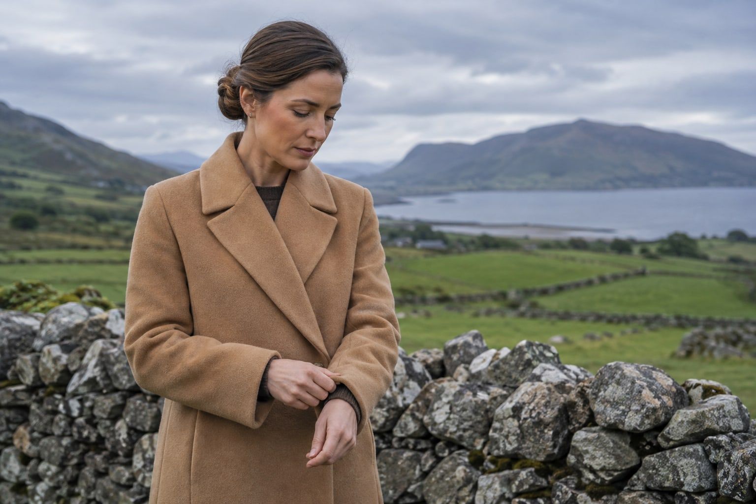 A woman in a camel wool coat adjusting her cuff beside a stone wall in green countryside.